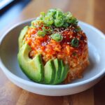A molded mound of spicy crab sushi bowl mixture served in a white bowl, garnished with sliced avocado, scallions, and sesame seeds.