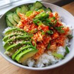 A close-up of a spicy crab sushi roll bowl featuring spicy shredded crab mix over rice, topped with sliced avocado, cucumber, and green onions.