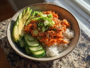 A close-up of a spicy crab sushi roll bowl featuring rice, bright orange spicy crab mix, sliced avocado, and cucumber.