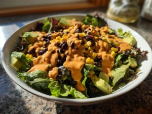 A vibrant bowl of spicy southwest salad featuring mixed greens, black beans, corn, avocado, and a generous drizzle of orange dressing.