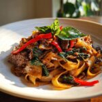 A close-up of glossy, wide rice noodles tossed with beef, basil, and bright red chili peppers in thai drunken noodles with beef.
