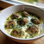 Close-up of a white bowl filled with swedish meatball soup, featuring browned meatballs in a creamy broth topped with fresh dill.