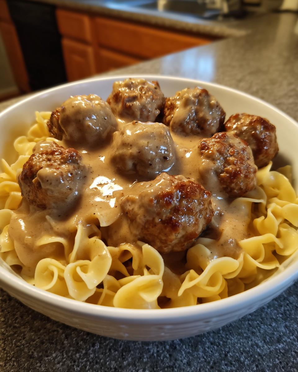 A close-up of swedish meatballs over egg noodles, covered in creamy brown gravy, served in a white bowl.