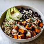 A close-up of a vibrant sweet potato buddha bowl featuring roasted sweet potatoes, quinoa, black beans, avocado, and tahini dressing.