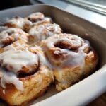 Close-up of gooey, freshly baked tiktok cinnamon rolls rhodes topped with thick white icing in a white baking dish.