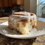 A single, freshly baked tiktok cinnamon rolls topped with thick white icing, sitting on a white plate.
