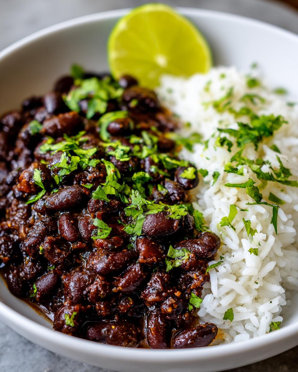 A close-up of a white bowl filled with rich black beans and rice, garnished with cilantro and a lime wedge.