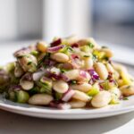 A close-up, bright photo of a mound of white bean salad mixed with chopped red onion and celery on a white plate.