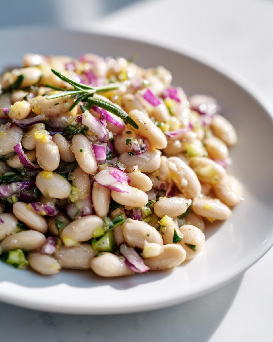 Close-up of a bright white bean salad mixed with diced red onion and herbs, garnished with a sprig of rosemary.