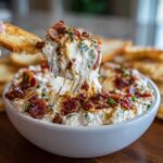 A piece of toasted bread is dipping into a bowl of creamy, bacon-topped cream cheese dip, showing a satisfying cheese pull.