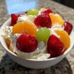 A close-up of a serving of cheesecake fruit salad topped with fresh strawberries, mandarin oranges, and green grapes over whipped cream.