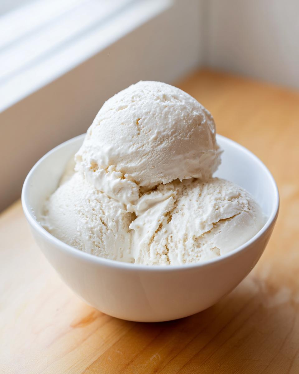 Two scoops of creamy, vanilla-colored cottage cheese ice cream served in a white bowl on a wooden surface.