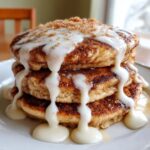 Close-up of a stack of fluffy cinnamon roll pancakes dripping with thick white icing and sprinkled with brown sugar.