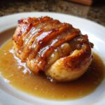 A close-up, mouthwatering shot of a baked apple dumpling covered in a thick, glossy caramel sauce, resting in a pool of sauce on a white plate.