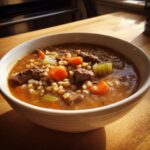 A close-up of a white bowl filled with rich beef barley soup, showing chunks of beef, barley, carrots, and celery.