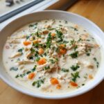 Close-up of a white bowl filled with homemade cream of chicken soup, featuring shredded chicken, carrots, and parsley.