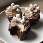 Three decadent hot chocolate cupcakes on a white plate, one with a bite taken out, topped with chocolate frosting and mini marshmallows.