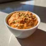 A close-up of a small white bowl filled with thick, orange-hued remoulade sauce, garnished with fresh snipped chives.