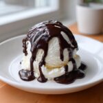 A scoop of vanilla ice cream generously covered in glossy, homemade chocolate sauce, served on a white plate.