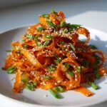 A pile of bright orange raw carrot salad ribbons topped with toasted sesame seeds and chopped green onions.